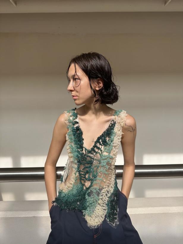 Eine junge Frau mit nassem Haar trägt ein aufwendiges, transparentes Oberteil in Grün- und Beigetönen.