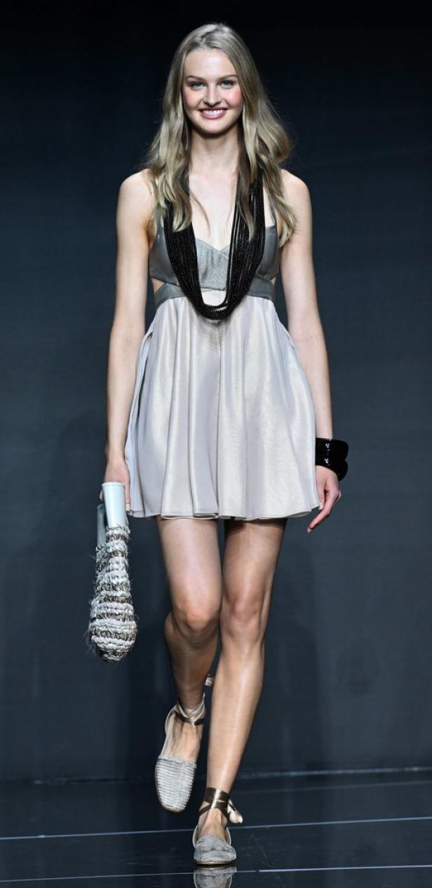 Ein Model präsentiert ein kurzes, helles Kleid mit auffälliger Halskette und Armreif auf dem Laufsteg.