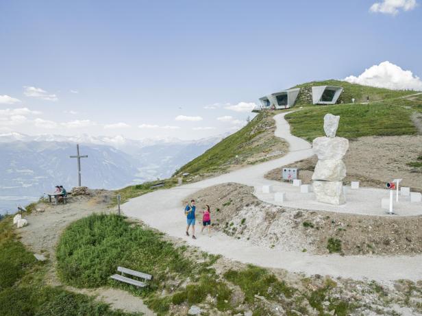 Ein Paar wandert auf einem Pfad auf einem Berg mit moderner Architektur im Hintergrund.