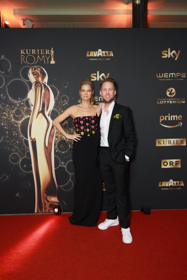 Ein Mann und eine Frau posieren vor einer Wand mit Logos für die „Kurier Romy“-Preisverleihung.