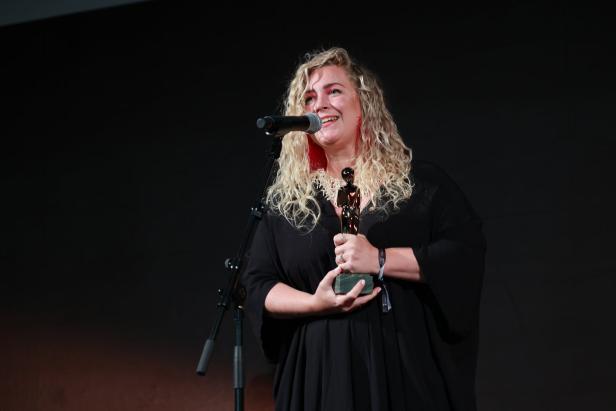 Eine Frau mit blonden Locken hält eine Auszeichnung in der Hand und lächelt in ein Mikrofon.