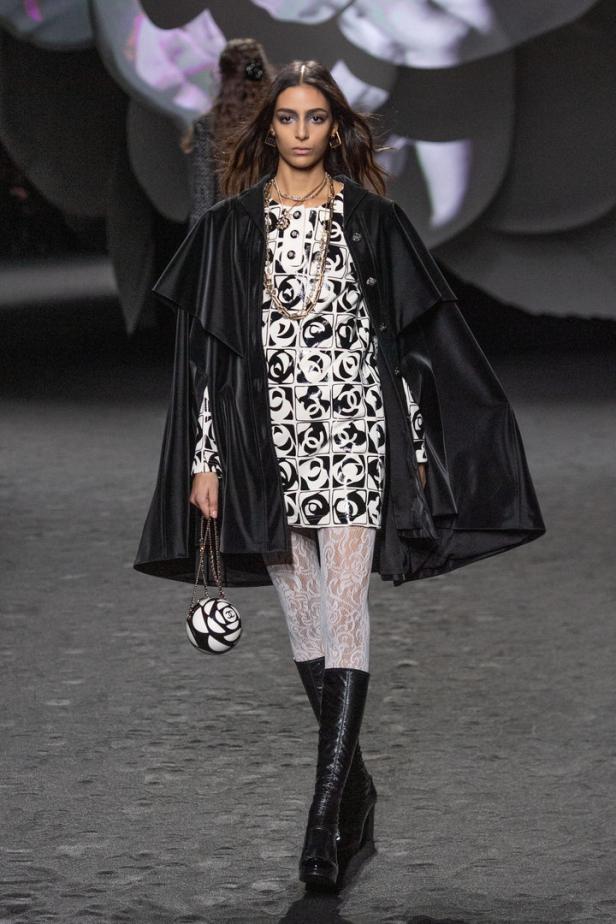 Ein Model präsentiert ein Kleid mit Chanel-Logo, eine schwarze Cape und passende Accessoires.