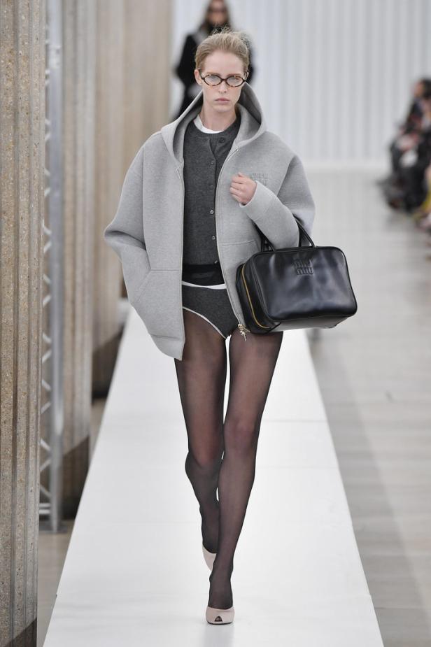 Eine Frau mit Brille und grauer Jacke läuft mit einer schwarzen Miu Miu Tasche über einen Laufsteg.