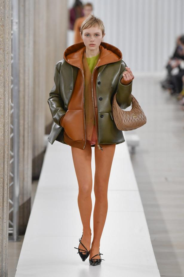 Eine Frau in einem grünen Ledermantel mit Kapuze und einer braunen Miu Miu Handtasche auf dem Laufsteg.
