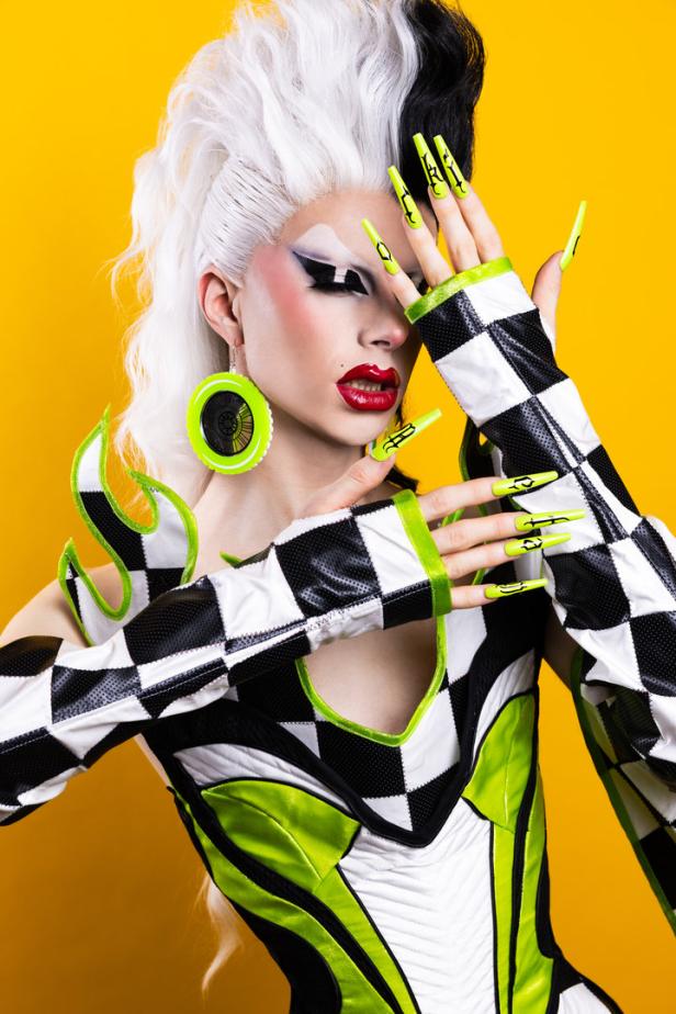 Eine Dragqueen mit schwarz-weißer Perücke, Schachbrettmuster-Outfit und grünen Akzenten posiert vor gelbem Hintergrund.