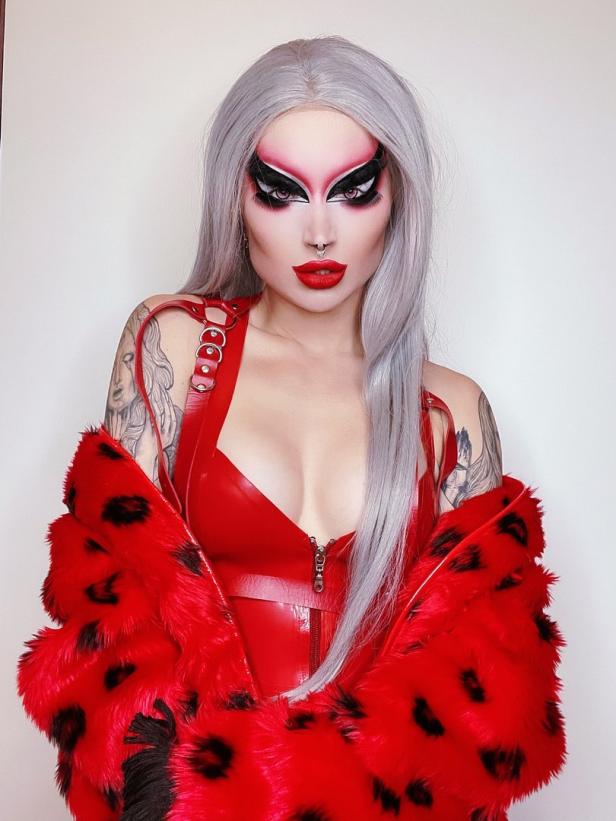 Eine Dragqueen mit silbernen Haaren, rotem Outfit und auffälligem Make-up posiert vor einer weißen Wand.