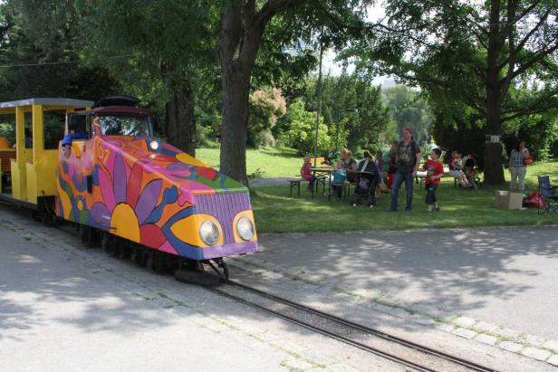 Eine bunte Parkeisenbahn fährt an einer Gruppe von Menschen vorbei.