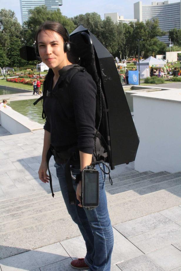 Eine Frau mit Kopfhörern trägt einen schwarzen, pyramidenförmigen Rucksack und ein Gerät am Handgelenk.