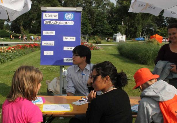 Personen sitzen an einem Tisch vor einem Schild der „Kleinen Sprachschule“.