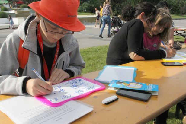 Ein Junge mit Hut schreibt auf eine kleine Tafel, während ein Mädchen daneben auf eine andere Tafel schreibt.