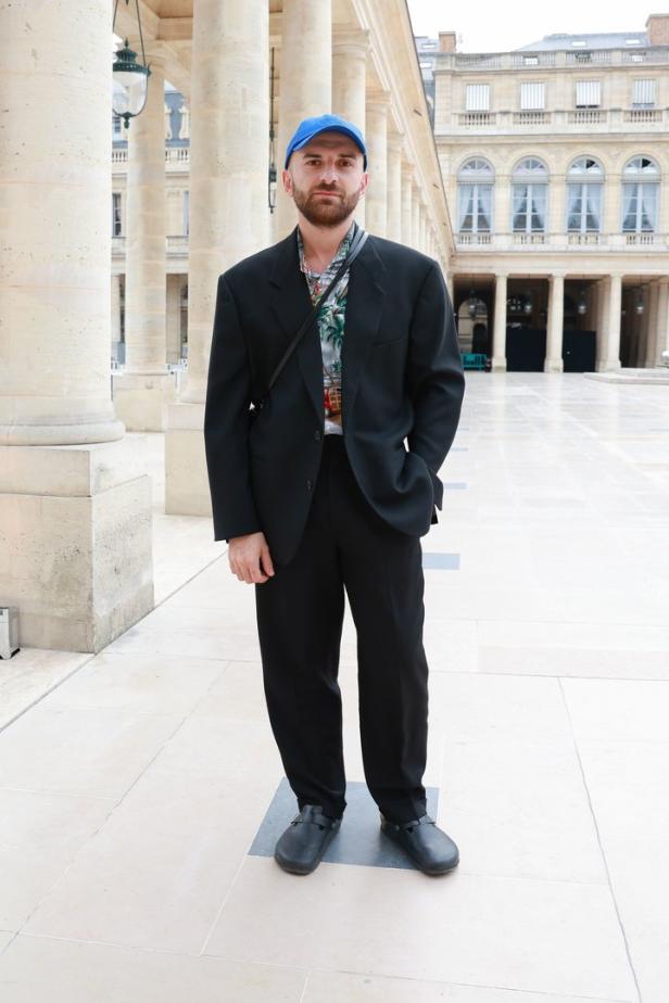 Ein Mann mit blauem Cap, schwarzem Anzug und Clogs steht im Freien.