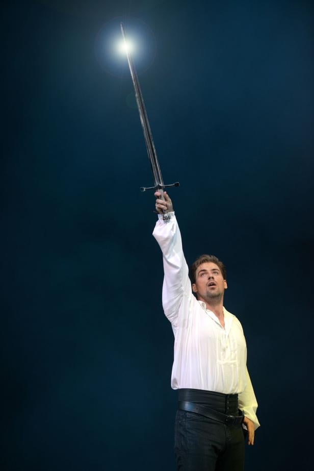 Ein Mann im weißen Hemd hält ein Schwert in die Höhe.