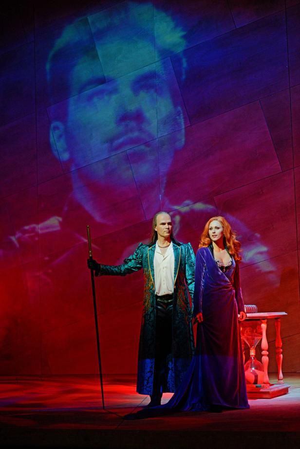 Zwei Schauspieler stehen auf einer Bühne vor einer großen Projektion eines Gesichts.