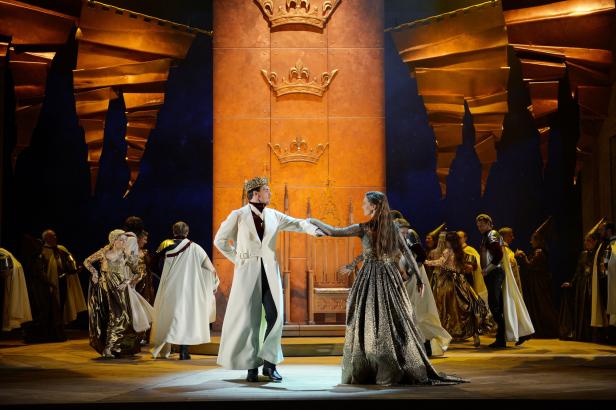 Auf einer Bühne stehen ein König und eine Königin in prächtigen Gewändern.