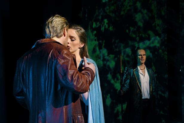 Eine Szene aus einem Theaterstück mit zwei Liebenden und einer mysteriösen Figur im Hintergrund.