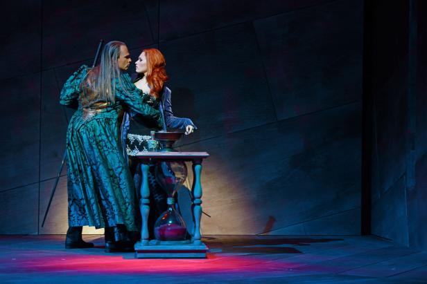 Auf einer Bühne stehen ein Mann und eine Frau an einem Tisch mit einer Sanduhr.