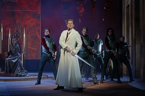 Ein Mann in einem weißen Mantel hält ein Schwert, umgeben von Männern in Rüstungen auf einer Bühne.
