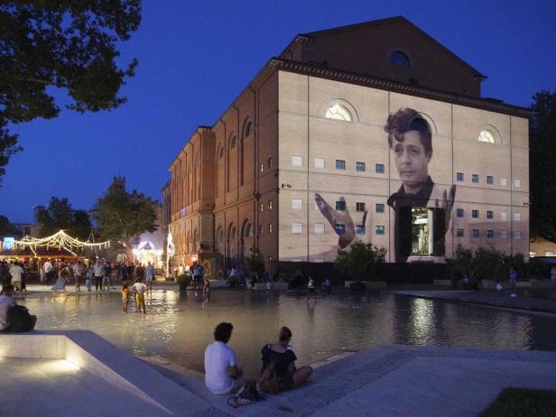 Am Abend wird ein Porträt auf die Fassade eines Gebäudes in der Nähe eines belebten Platzes projiziert.