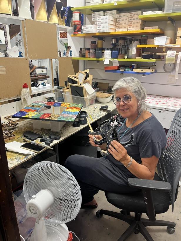 Eine Frau sitzt in einem Atelier und hält ein Mosaik-Kunstwerk in den Händen.