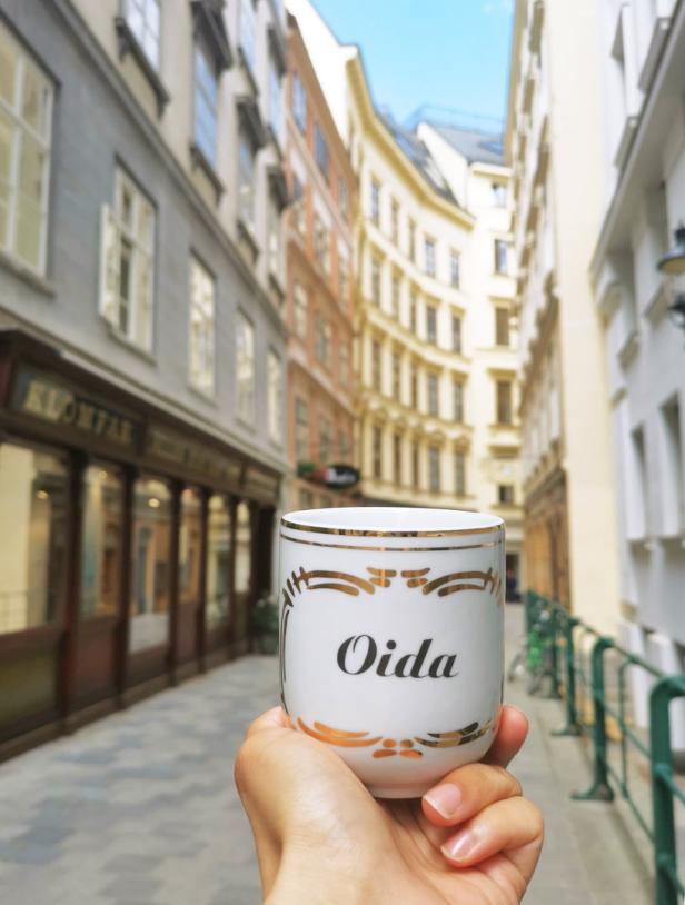 Eine Hand hält eine Tasse mit der Aufschrift „Oida“ in einer Wiener Gasse.