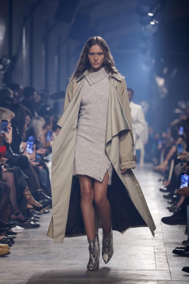 Eine Frau in einem beigen Trenchcoat und einem grauen Kleid auf einem Laufsteg.