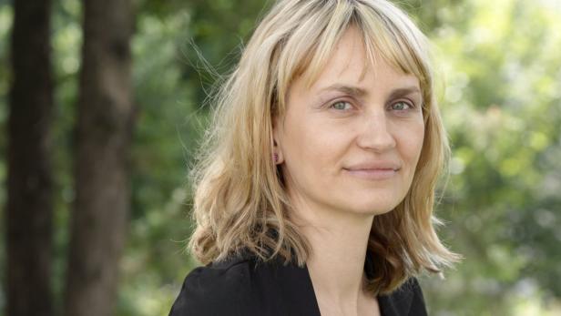 Porträt einer Frau mit blonden, gewellten Haaren und einem schwarzen Blazer vor einem grünen Hintergrund.