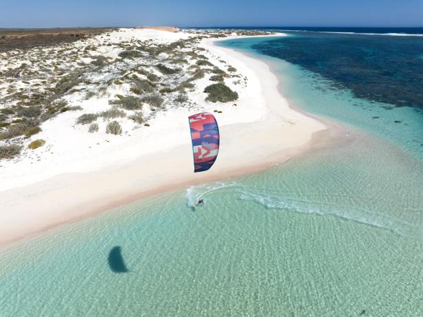 Ein Kitesurfer fährt auf einer Sandbank entlang, mit türkisfarbenem Wasser im Hintergrund.