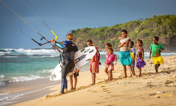 Eine Kitesurferin steht am Strand, beobachtet von einer Gruppe einheimischer Kinder.