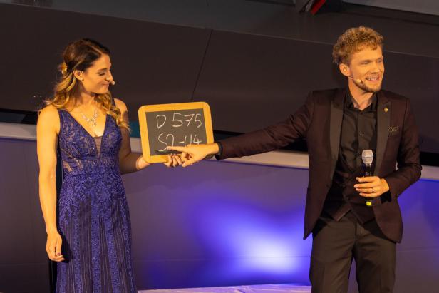 Eine Frau im Abendkleid und ein Mann im Anzug präsentieren eine Tafel mit der Aufschrift „D 575 S2-114“.