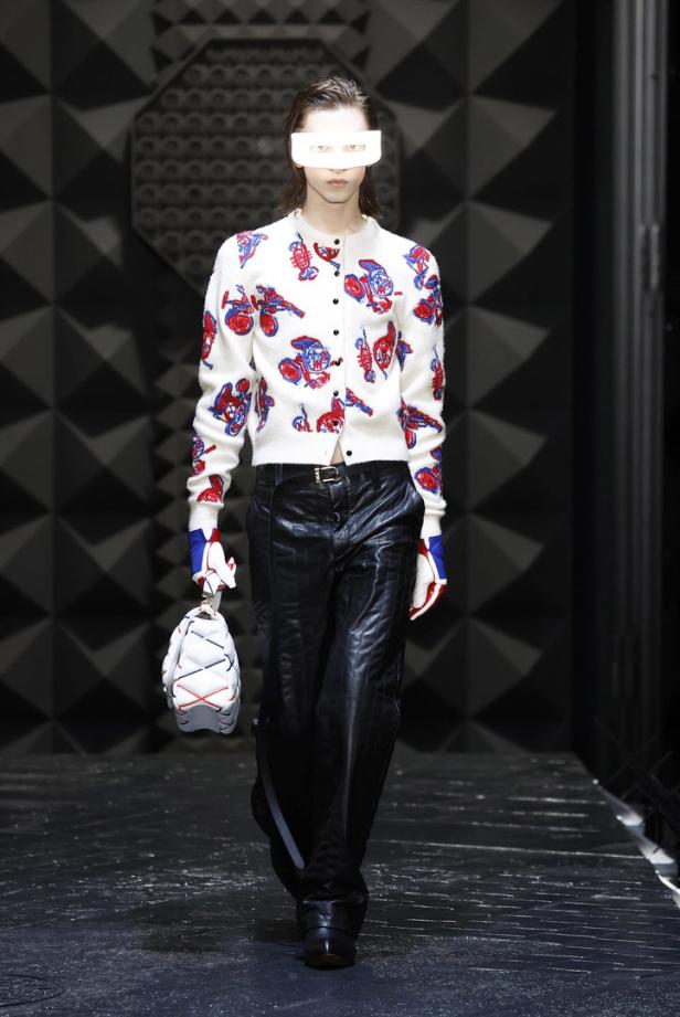 Ein Model mit weißer Augenmaske auf dem Laufsteg, gekleidet in einen Cardigan mit Cartoon-Motiven und glänzende Hosen.