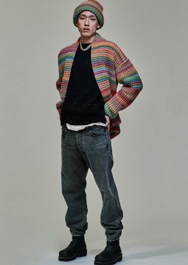 Ein Mann mit Mütze und Strickjacke in Regenbogenfarben posiert selbstbewusst.