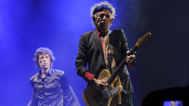 Keith Richards spielt Gitarre auf der Bühne, während Mick Jagger im Hintergrund zu sehen ist.