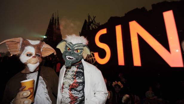 Zwei Personen mit Gremlins-Masken stehen vor einer großen Leuchtreklame mit der Aufschrift „SIN“.