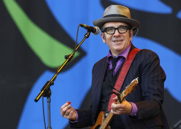 Elvis Costello tritt mit Hut und Gitarre auf einer Bühne auf.