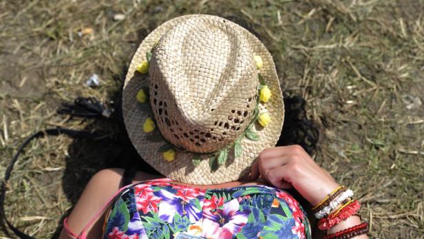 Eine Frau liegt im Gras, ihr Gesicht ist unter einem mit gelben Blumen geschmückten Strohhut verborgen.