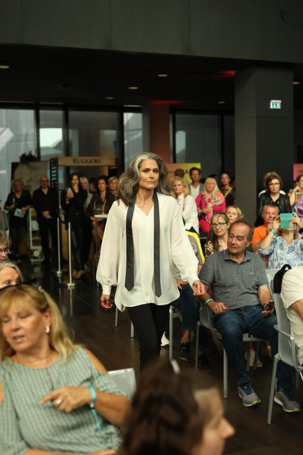 Eine Frau mit grauem Haar läuft auf einem Laufsteg vor Publikum.