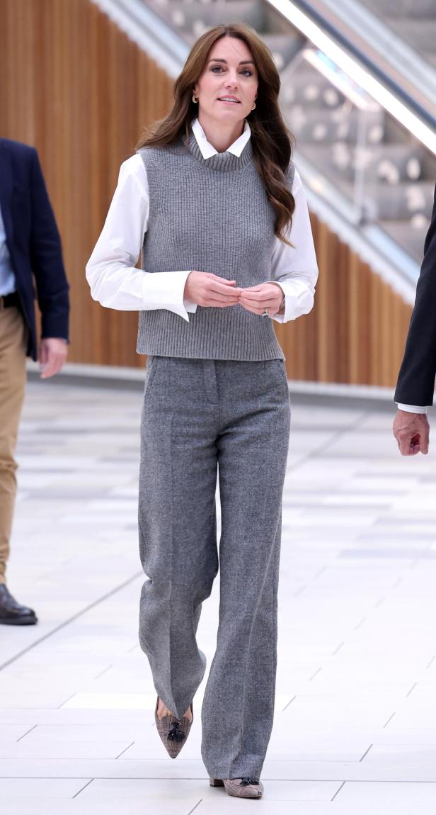 Prinzessin Kate mit grauer Anzughose und dazu passendem grauen Pollunder. Darunter trägt sie ein weißes Hemd und dazu spitze Schuhe.