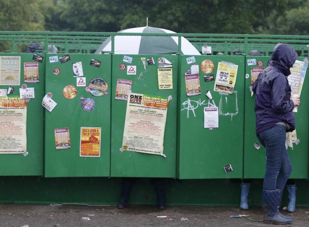 Eine Person steht im Regen vor einer grünen Wand voller Festivalplakate.