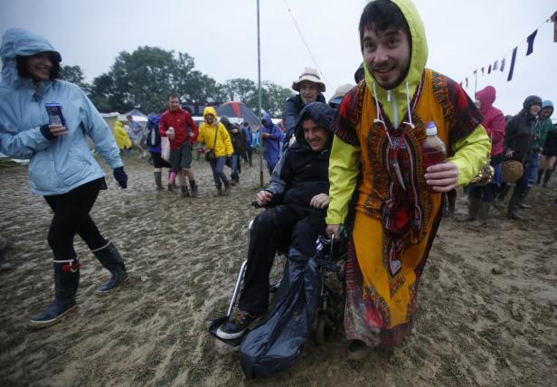 Eine Gruppe von Menschen schiebt eine Person im Rollstuhl durch den schlammigen Boden eines Festivals.