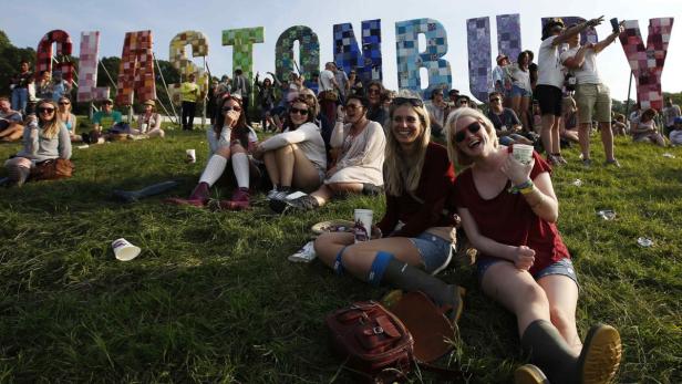 Besucher sitzen vor dem „Glastonbury“-Schriftzug auf dem Glastonbury Festival im Grünen.