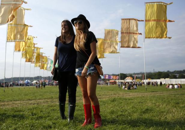 Zwei junge Frauen in Gummistiefeln auf einem Festivalgelände mit gelben Fahnen im Hintergrund.