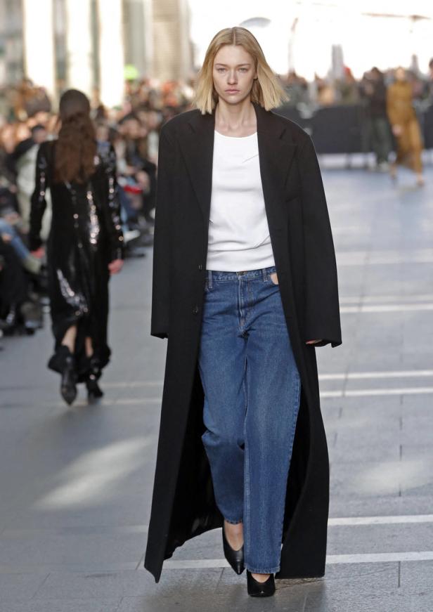 Eine Frau mit blonden Haaren trägt einen langen schwarzen Mantel, Jeans und schwarze Schuhe.