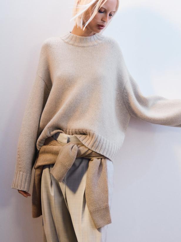 Eine Frau mit blonden Haaren trägt einen beigen Pullover und eine helle Hose.
