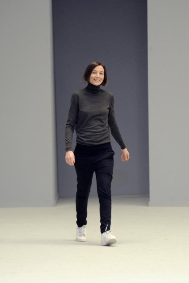 Eine Frau mit dunklen Haaren, gekleidet in einen Rollkragenpullover und dunkle Hose, geht auf einem Laufsteg.