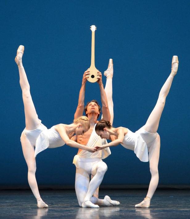 Ein Ballett-Tänzer kniet mit einer Laute in den Händen, flankiert von zwei Tänzerinnen in weißen Kostümen.