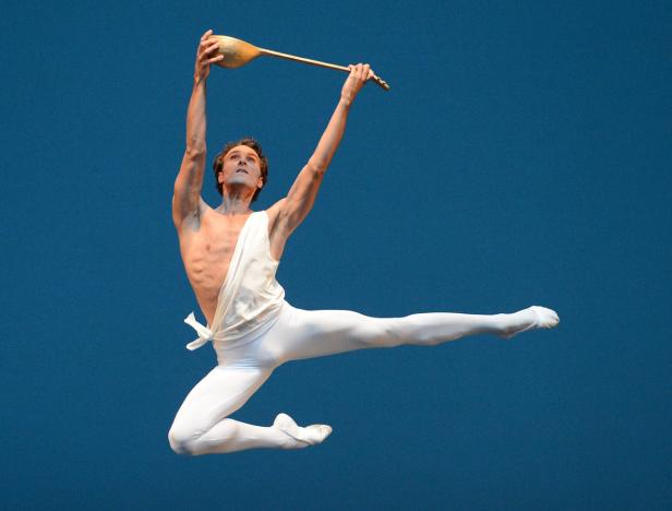 Ein männlicher Balletttänzer springt mit einem goldenen Gegenstand in der Hand vor einem blauen Hintergrund.