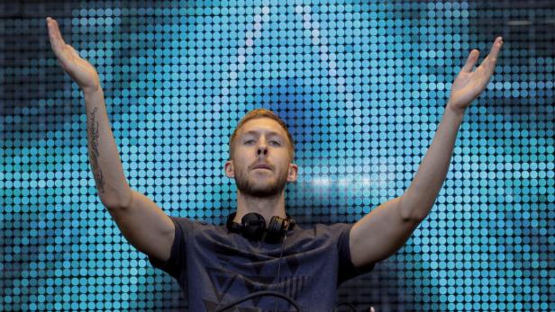Calvin Harris steht mit erhobenen Armen vor einer blau leuchtenden Wand.
