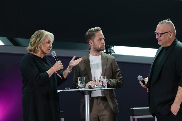 Drei Personen stehen auf einer Bühne und sprechen in Mikrofone.