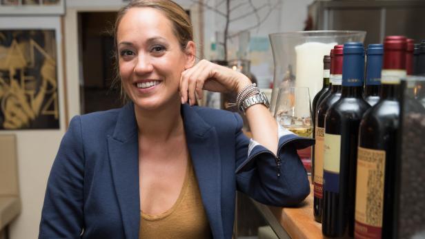 Eine lächelnde Frau sitzt an einer Bar, neben ihr stehen Weinflaschen.