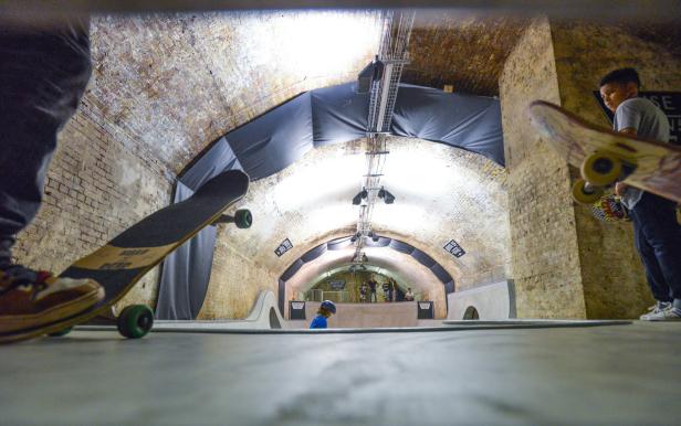 Kinder und Erwachsene in einem Skatepark in einem Tunnel mit Ziegelwänden.